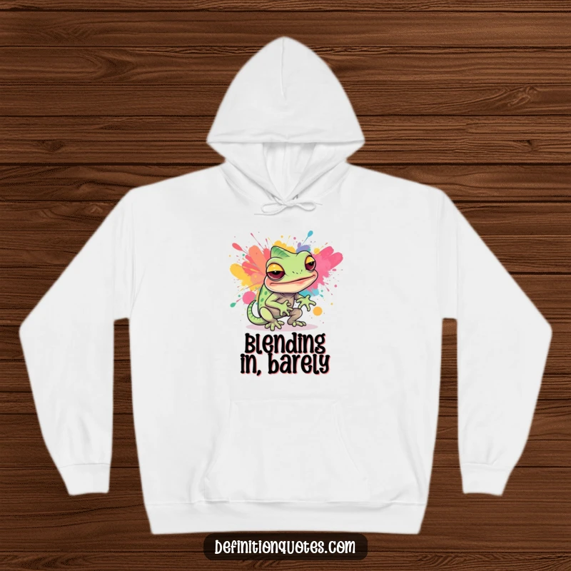 Funny, cozy hoodie featuring a sly chameleon character attempting to blend into a vibrant abstract paint splatter, showcasing artistic camouflage.